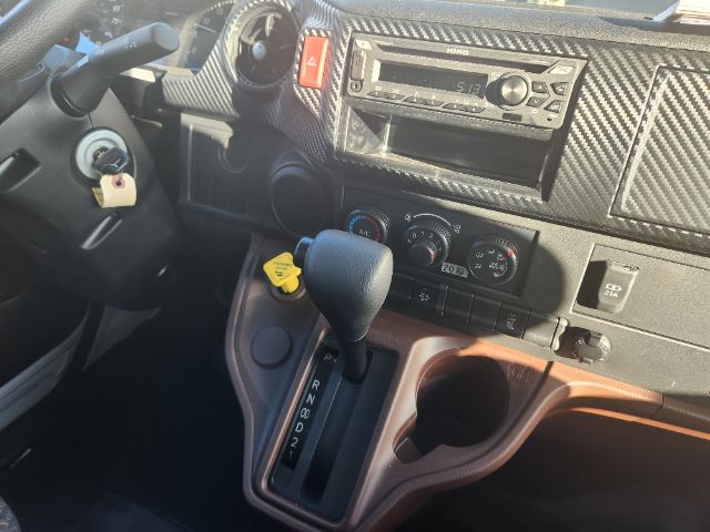 Interior of a 2025 Hino L6 cab with automatic transmission, gear selector PRND321, steering column key, dashboard controls, radio, and climate dials.