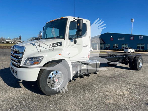 Front three-quarter view of a white 2025 Hino L6 cab and chassis truck, highlighting the cab, chrome grille, large windshield, and exposed dual rear axles with a flatbed chassis.