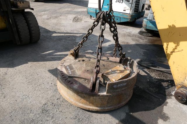 Circular metal debris with chains lifting lug attached, in an outdoor industrial yard with machinery in background.