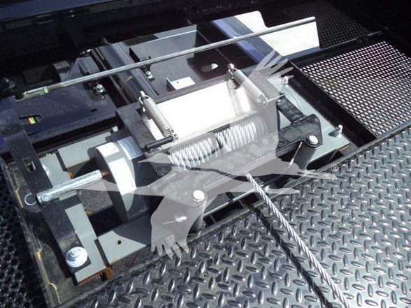 Tractor-trailer winch assembly with steel cable and drum on diamond plate platform, mounted under truck bedside. Visible are springs, cables, and mounting brackets.
