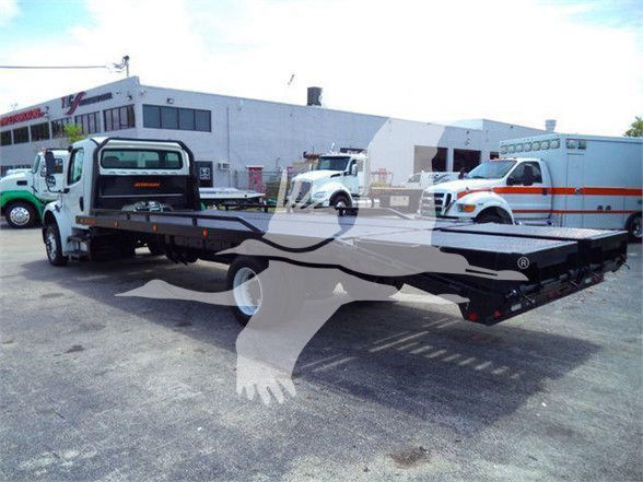 Flatbed tow truck with long black platform, Freightliner Business Class M2 106 style cab in background, showing a low-profile flatbed carrier.