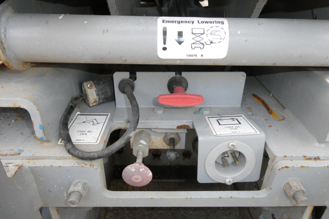 Genie GS1930 scissor lift fuel/electrical panel area showing 115V AC 15A receptacle and 115V 12A receptacle with cords and red hand crank. Emergency lowering label on guard rail.