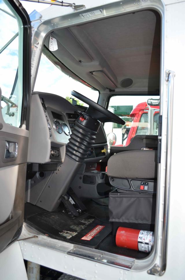 Truck cab interior of a 2014 Kenworth T370 with steering wheel, dashboard gauges, and doorway; Floor mat with red label; Fire extinguisher visible under seat.
