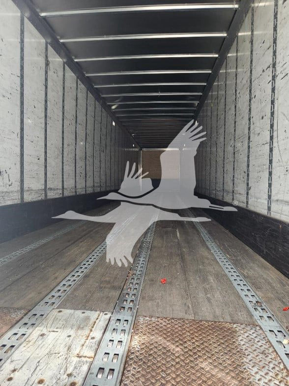 Interior of a long trailer with metal sidewalls and a wooden floor, empty, viewed from the back toward the front. A translucent bird graphic overlay sits across the scene.
