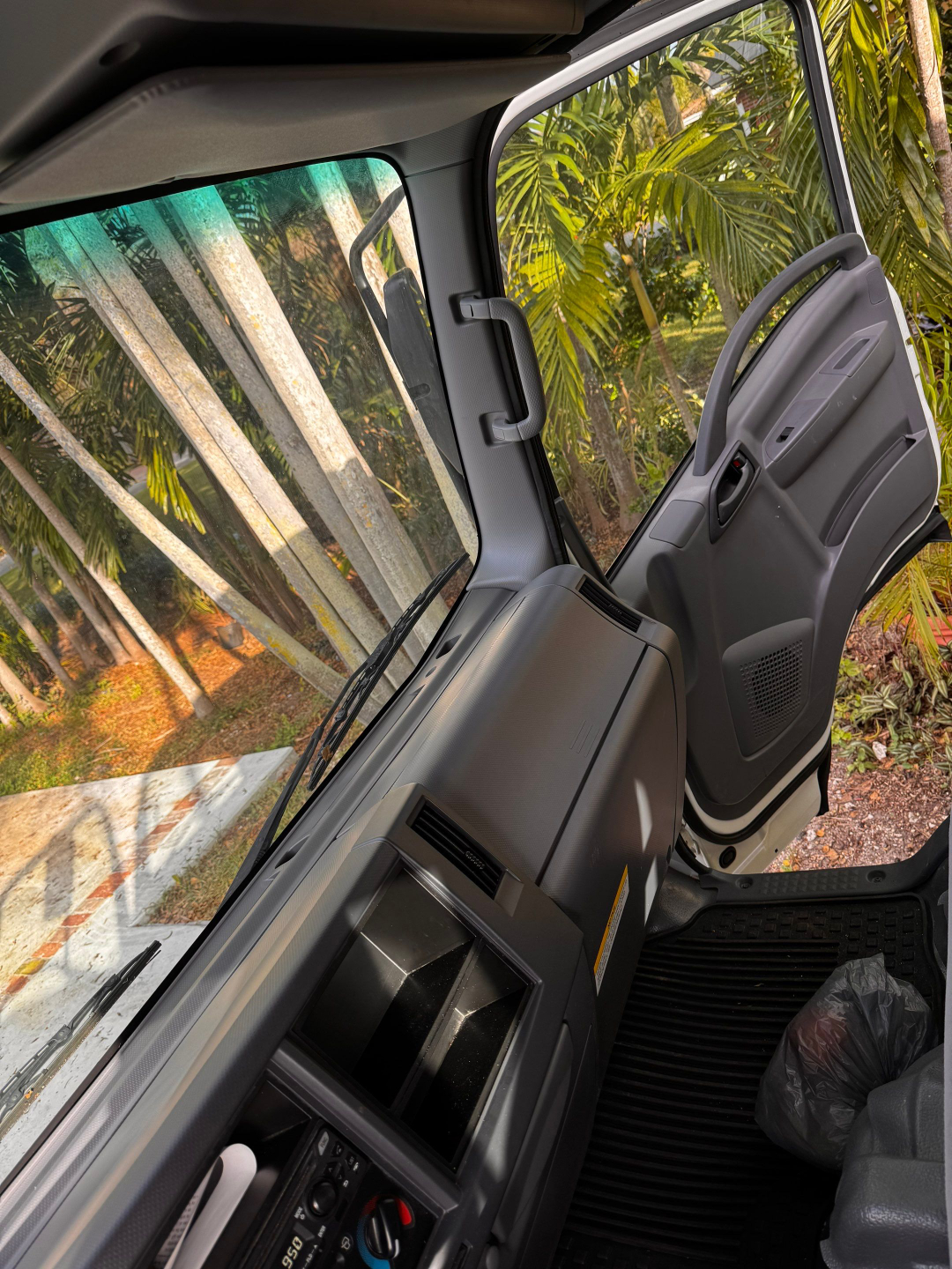 Isuzu NQR cab interior showing driver’s door, instrument panel, and windshield with palm trees outside.