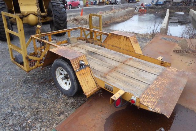 Trailer deck with ramps and yellow frame, single axle, wooden planks on bed, visible rust accents, set on a muddy trailer lot.