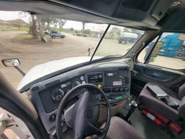 Vehicle cockpit of a Freightliner Cascadia truck showing steering wheel, dashboard with gauges, and center console; windshield view of yard with other trucks.