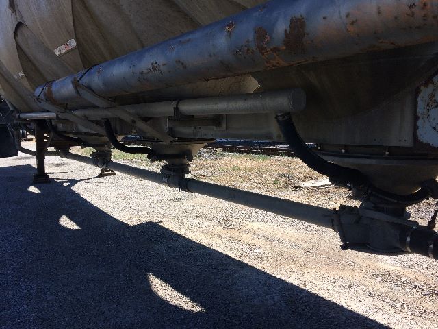 Trailer undercarriage showing a rust-stained cylindrical tank with plumbing, valves, and support beams; hydraulic-looking pipelines and fittings along the bottom.
