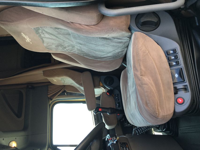 Truck cabin interior showing driver and passenger seats with beige upholstery and control switches near seat bases inside a Freightliner Cascadia 125 truck.