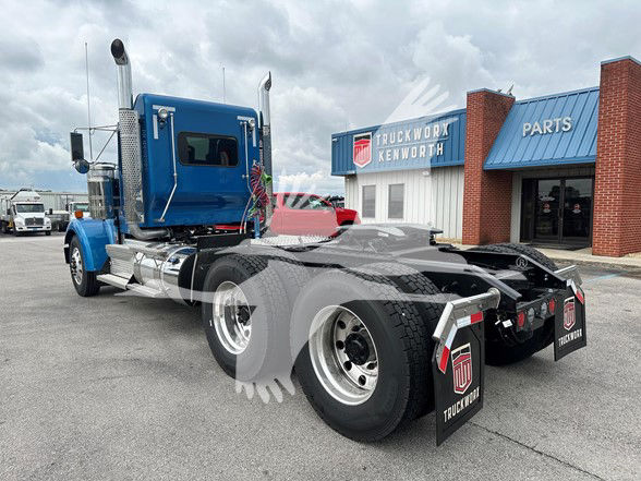 Kenworth W900L conventional daycab truck in blue, rear tractor with dual rear axles and mud flaps, dealership building in background.