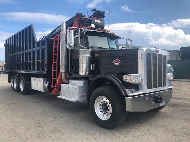 Peterbilt 388 dump truck with grapple system, black cab, chrome bumper, large multi-axle dump bed and crane on top, blue sky with clouds.