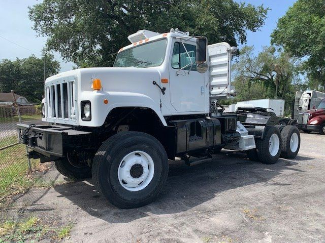 White International truck with sleeper/cab and dual rear axles, front grille, headlights, and heavy-duty chassis. Flatbed absent, showing front left wheel and steering; no visible branding text besides general truck features.