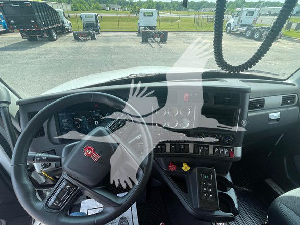 Kenworth T880 cockpit interior showing steering wheel with Kenworth emblem, instrument cluster, and dashboard controls. Windows reveal truck yard with other tractors.