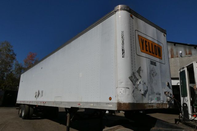 Trailer box side and front corner with a yellow YELLOW label on rear doors, generic white cargo trailer, visible riveted panels and wheel area.