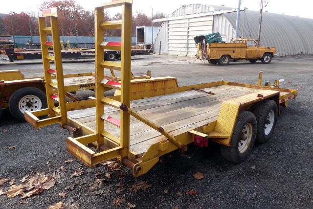 Industrial trailer with dual rear axles and wooden deck, yellow metal ramps on back, parked in lot with other equipment.