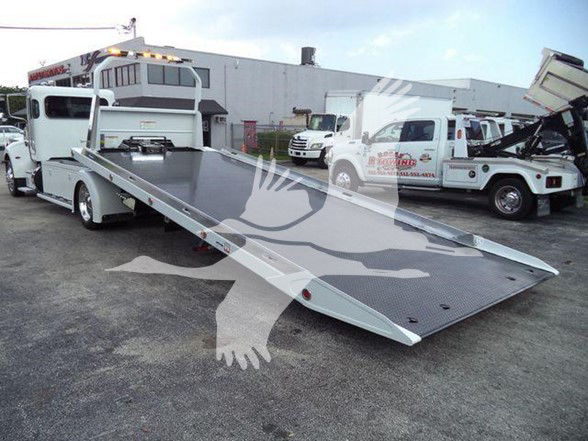 Truck with a lowered flatbed tow bed, Peterbilt cab visible in background, multiple tow trucks in lot.