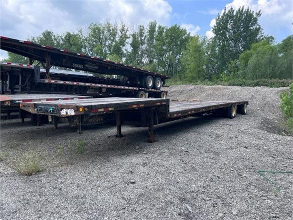 Dynaweld steel stepdeck trailer in a gravel lot, flatbed section extended with support legs visible, other trailers stacked nearby.