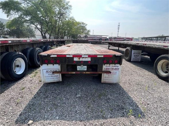 Flatbed trailer rear view showing multiple red circular taillights, mud flaps, and a wooden deck with white support blocks on gravel lot.