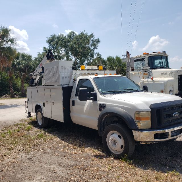 Ford F-550 utility truck with service body and equipment mounted in rear, white cab and utility compartments, side mirror, headlights visible.
