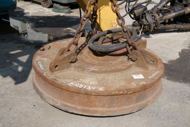 Circular iron weight with chain and lifting hooks attached, rusted surface, on concrete ground; industrial machinery background.