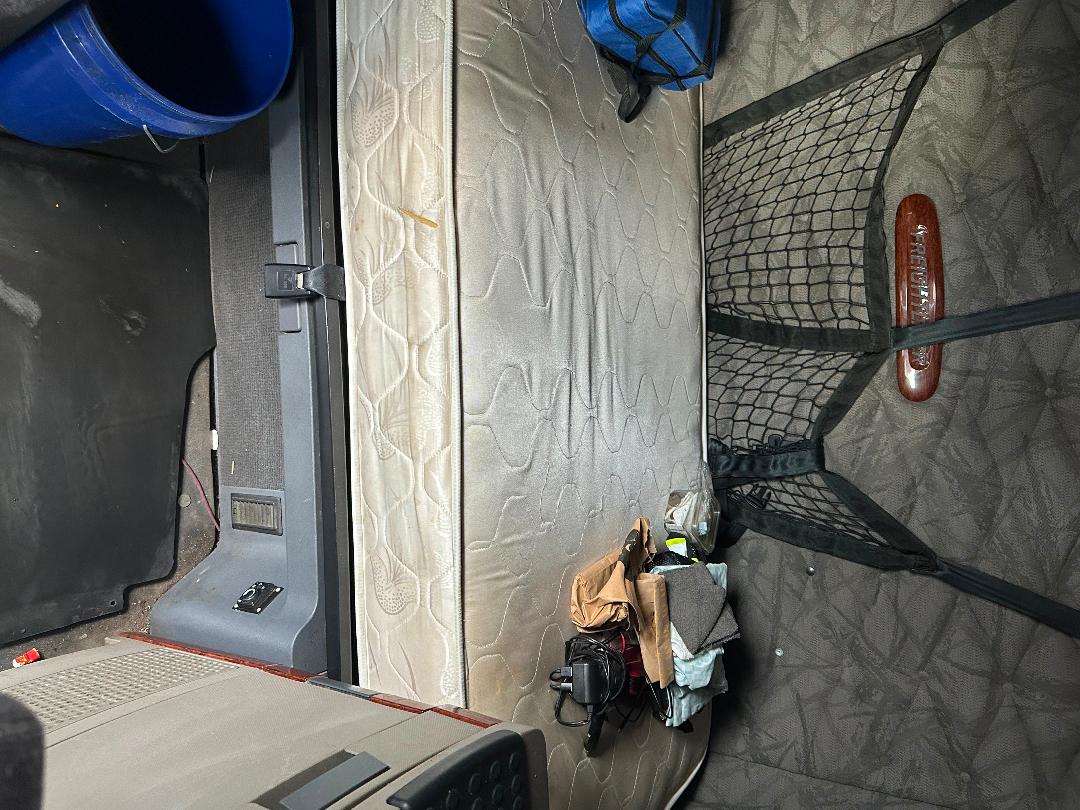 Cab sleeper area of a 2011 Freightliner Cascadia showing a mattress, net cargo bag, clutter near side wall, and a window shade handle.