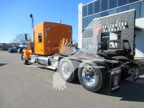 Kenworth W900L tractor unit with orange sleeper cab and chrome fuel tanks, parked outside a truck dealership.