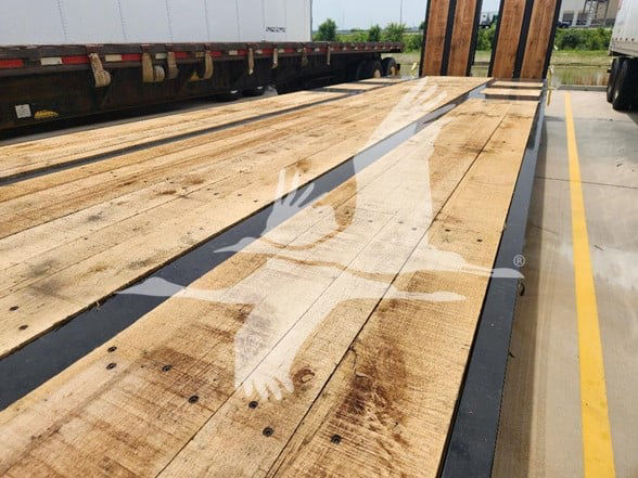 Flatbed trailer deck showing wooden planks with black edge rails and reflection on surface.