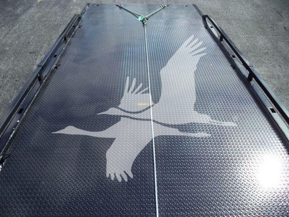 Truck bed with diamond plate surface featuring a large white bird graphic raising wings. Corner tie-down points visible along edges toward front. Center seam line and fasteners.