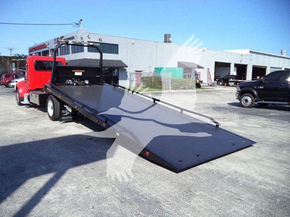 Peterbilt 337 tow truck with a tilted flatbed ramp in a lot, red cab, loading ramp extended, industrial buildings in background.