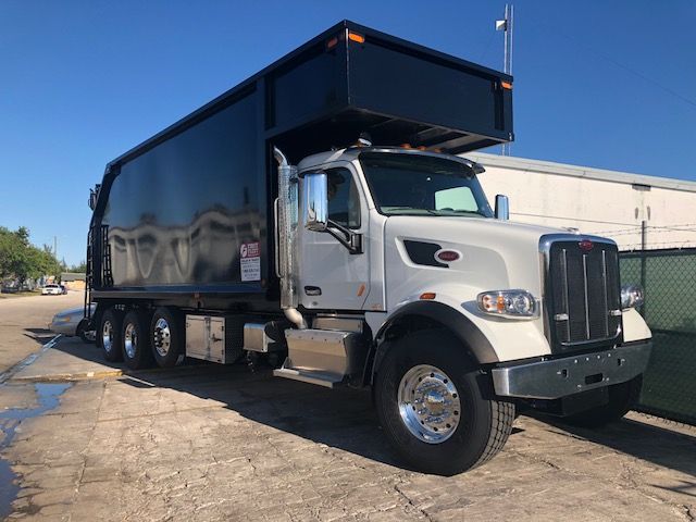 2018 Peterbilt 567 grapple truck with silver cab and large black dump trailer, chrome front bumper, and dual rear axles.