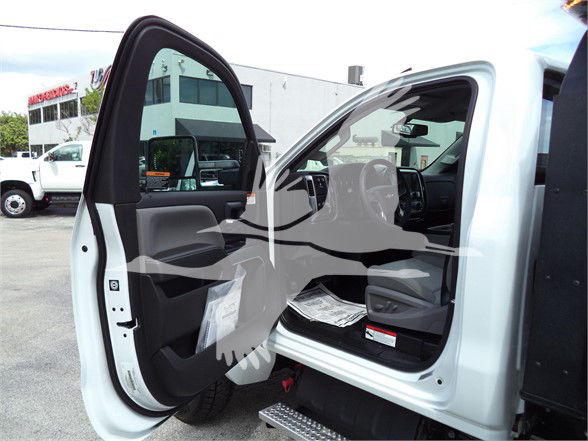 Truck cab interior visible with open driver door; dashboard, steering wheel, and front seats; light gray interior and white exterior of a Chevrolet Silverado 6500HD at a dealership lot.