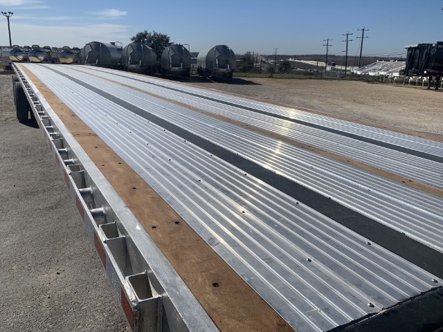 Long view of a 48ft flatbed trailer with metal corrugated decking and wooden accents, parked in an open lot with other trailers in the background.