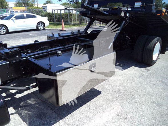 Truck chassis and bed frame showing a large metal tool/storage box and hydraulic components under the bed, with a white car in background.