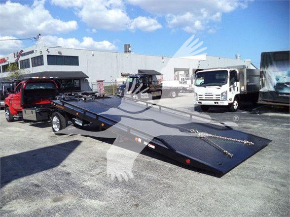 Tow truck flatbed/dolly with ramp surface, rear chains, and bumper lights in lot; background shows white trucks and a building.