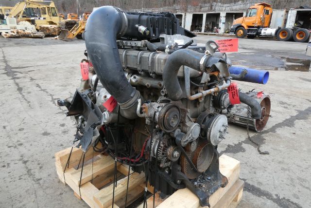 Engine core with multiple belts, hoses, and pulleys mounted on a wooden pallet. Visible components include a large black upper intake hose, grey valve covers, belt-driven pulleys, and red tags on some hoses.