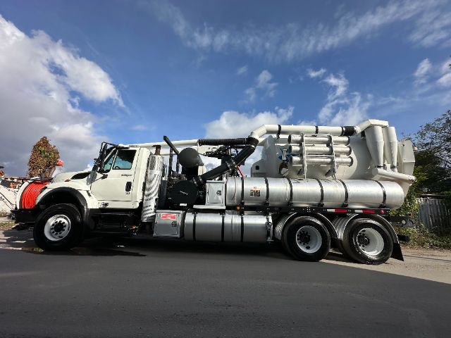 International 7500 vacuum truck side view showing large cylindrical vacuum tank with multiple hoses and exhaust components, white cab, dual rear axles, clear blue sky.
