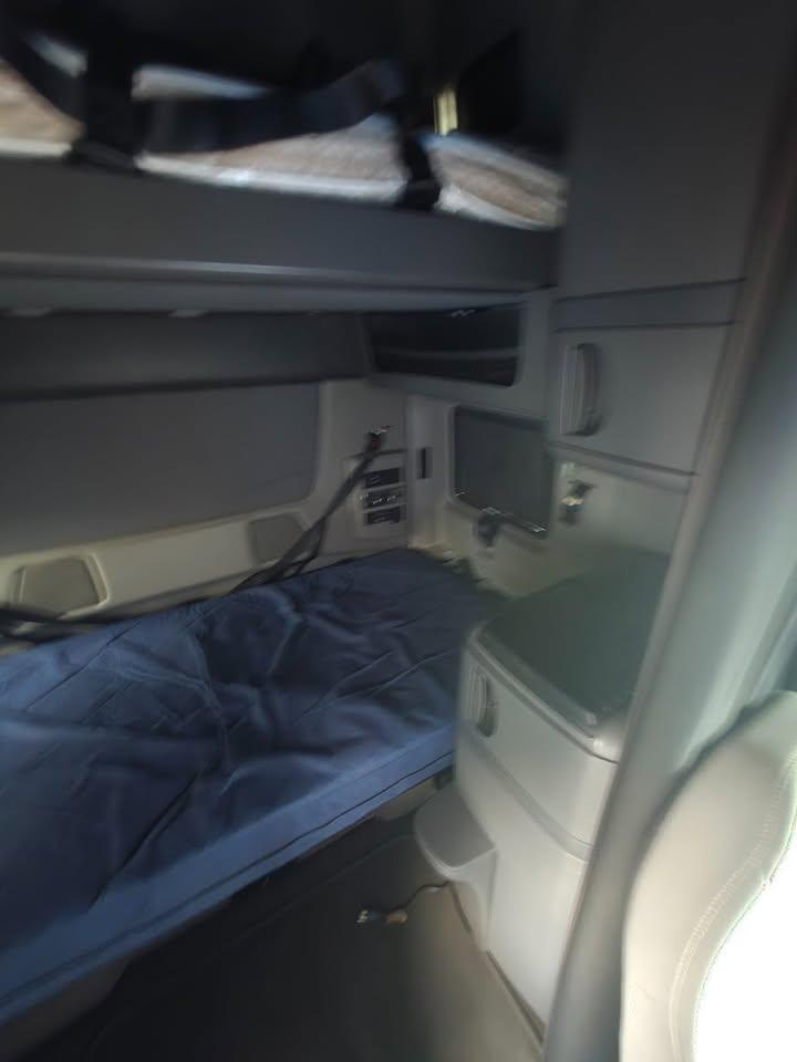 Truck cab interior showing bunk bed with blue blanket, dashboard controls, small monitor, storage cabinets.
