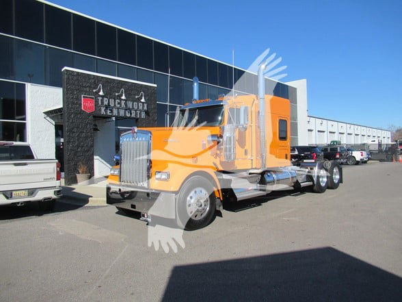 Orange 2025 Kenworth W900L conventional sleeper truck parked in lot with dealership building in background.