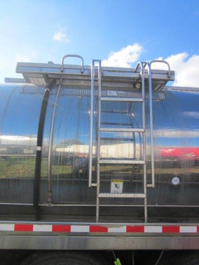 Tank truck side with ladder and manway access on polished stainless steel tank. Ladder attached, top access platform with safety rails, suction hose on left, gauge on right, safety sticker near ladder.