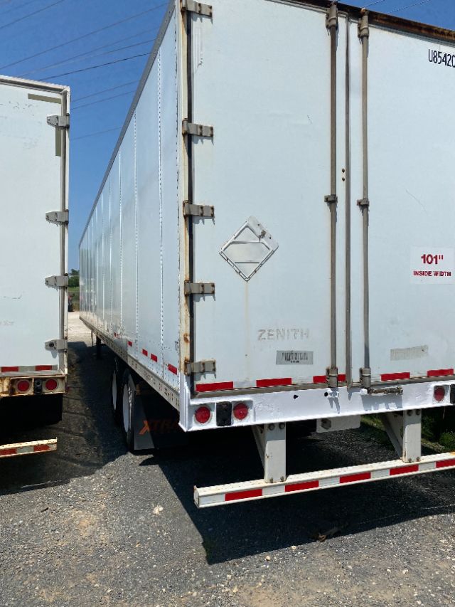 Rear view of a white Wabash Duraplate trailer with double rear doors, metal hinges, reflective tape, and rust spots. VIN/markings and safety decals visible.