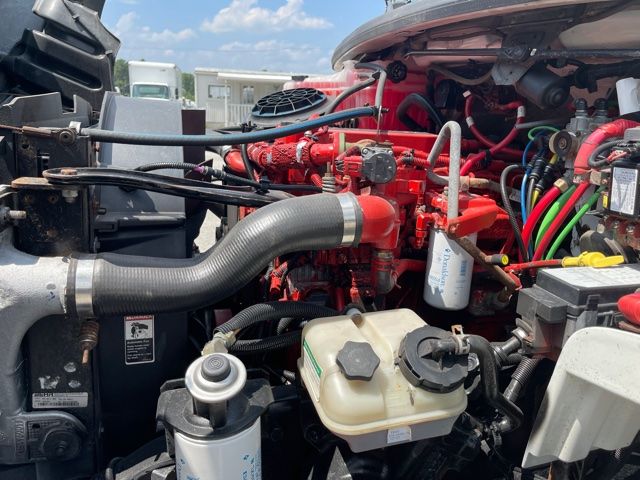 Engine bay of a 2013 Freightliner M2 with red engine components, hose assemblies, and fluid reservoirs visible.