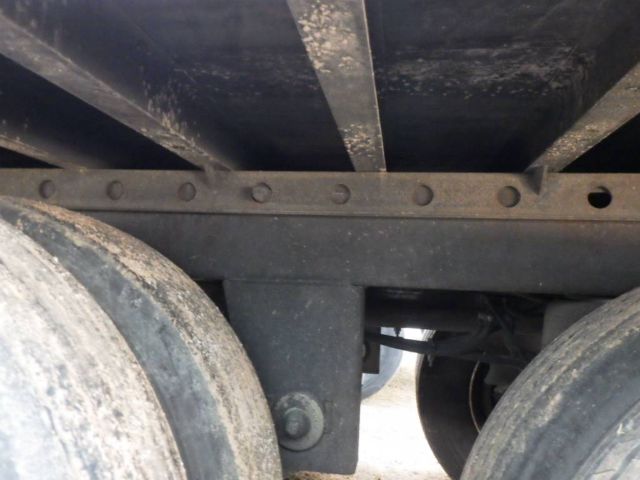 Underneath view of a utility trailer chassis showing wheels and frame with riveted crossmember; dirt and wear on metal surfaces.