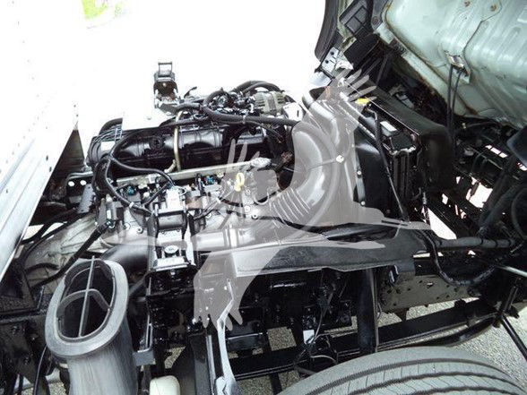 Isuzu NPR HD engine bay view with exposed components and front wheel, showing engine block, hoses, and fluid reservoir area.