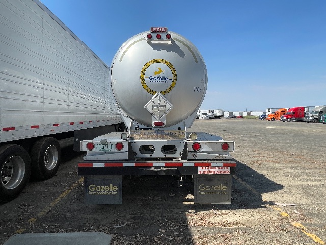 Back view of a silver cylindrical tanker trailer with Gazelle Transportation Inc. logo, taillights, warning placards, and supporting mud flaps in a lot with other trailers.