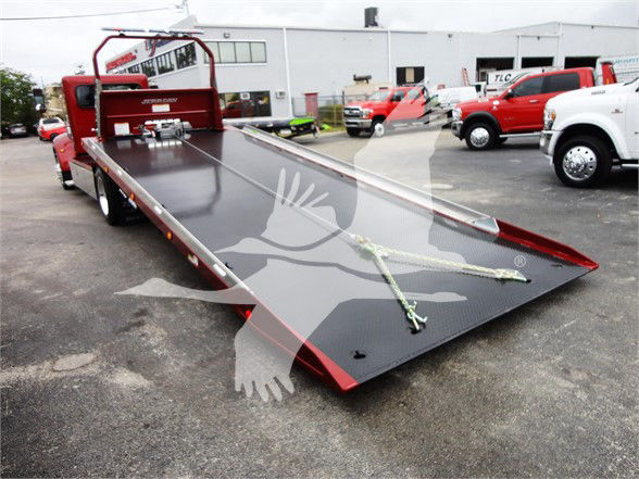 Flatbed tow truck bed on a 2022 Peterbilt 337 with a metal ramp and tiedowns in a dealership lot.