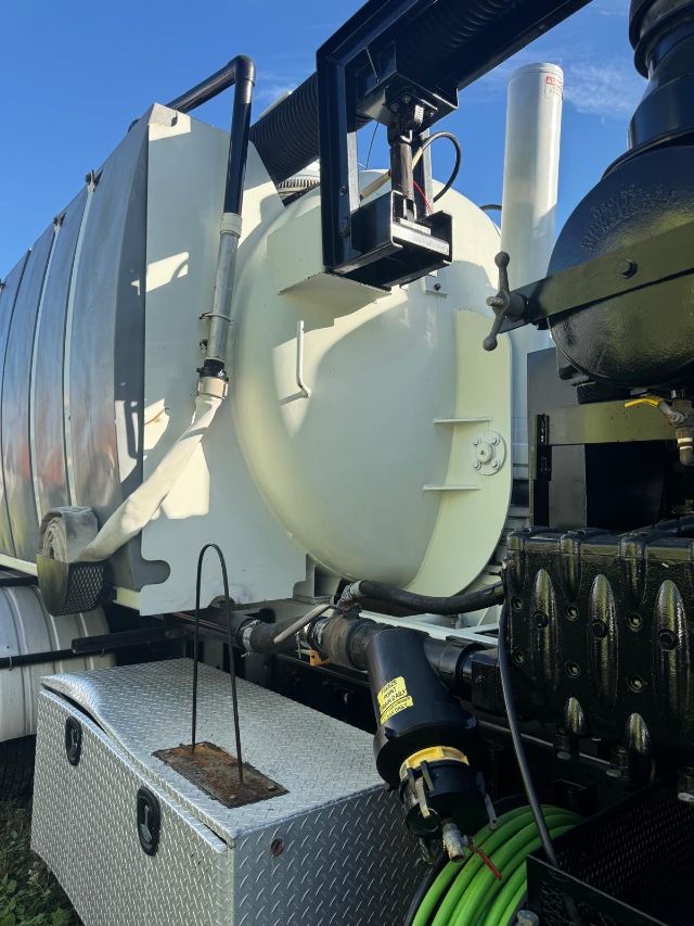 Front view of a 2014 Freightliner 114SD vacuum truck shows a large white cylindrical tank, hose reel, and diamond-plate toolbox with a green hose coiled nearby.