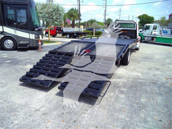 Flatbed tow truck in yard with dual foldable ramps extended, Freightliner Business Class M2 106 style cab visible in background.