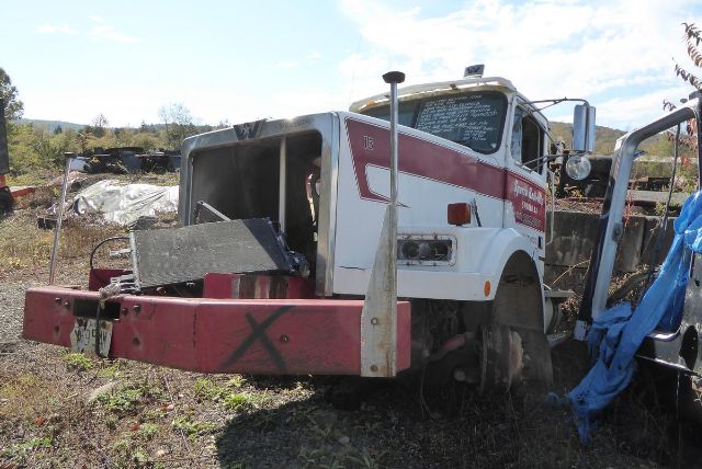 Cabover truck in salvage yard with red front bumper and white cab, missing front section, weathered surroundings.