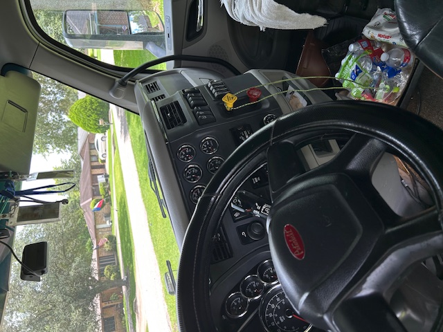 Peterbilt truck interior dashboard and steering wheel, with gauges and ignition area visible. Various controls and a yellow tag hang from a cord near the center. Plastic bottles and items on the floor. Side window and windshield view of a r