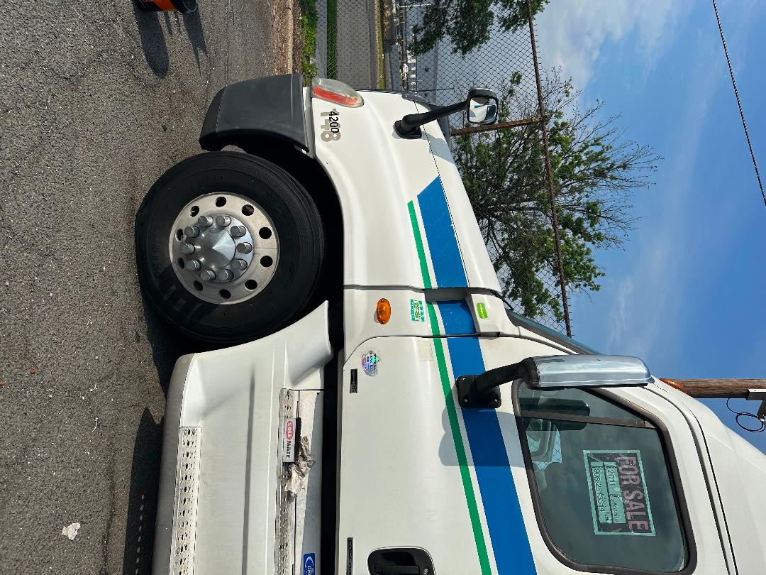 Truck side profile showing front wheel, cab with blue and green stripe graphics, Freightliner logo, side mirror, and cab door; visible 'FOR SALE' sign in window.